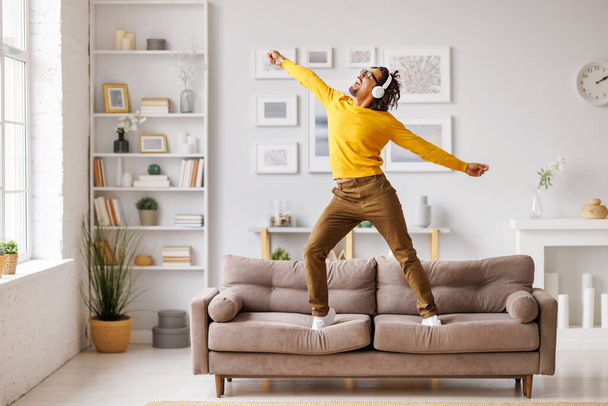 Full body uomo nero in abiti casual e cuffie wireless alzare le braccia, saltare e ballare sul divano divertendosi in soggiorno nel fine settimana a casa - Foto, immagini