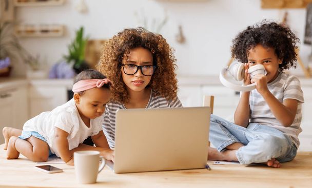 Jonge geïrriteerde gestresste Afro-Amerikaanse moeder bedekt oren en kijkt naar laptop scherm terwijl het werken op afstand van huis met twee kleine kinderen, wordt afgeleid van het werk door lawaaierige stoute kinderen - Foto, afbeelding