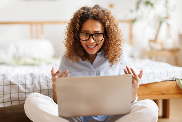 expresiva emocional afroamericana adolescente sentada cerca de la cama en casa sonriendo y gesticulando durante una videollamada a un amigo a través de un ordenador portátil con expresión excitada - Foto, imagen