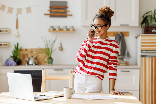 Jeune femme afro-américaine souriante et joyeuse dans des lunettes parlant au téléphone et regardant l'écran d'ordinateur portable tout en travaillant à distance dans un bureau à domicile confortable dans la cuisine moderne légère  - Photo, image
