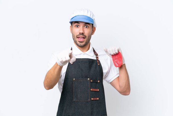 Homem caucasiano açougueiro vestindo um avental e servindo carne cortada fresca sobre fundo branco isolado surpreso e apontando frente - Foto, Imagem