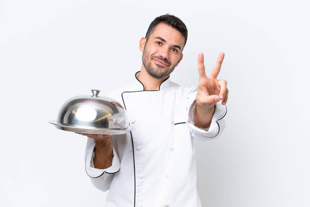 Молодий шеф-кухар з підносом ізольовані на білому тлі посміхаючись і показуючи знак перемоги
 - Фото, зображення