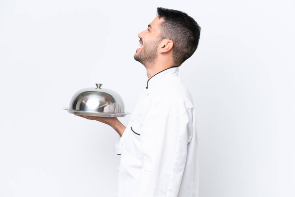 Jonge chef-kok met dienblad geïsoleerd op witte achtergrond lachen in laterale positie - Foto, afbeelding