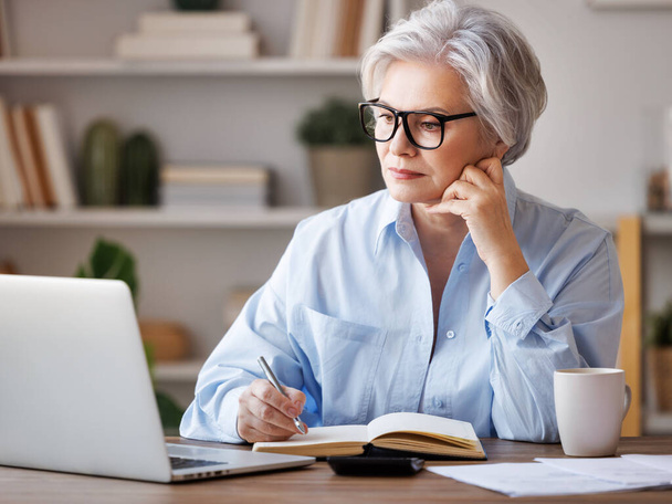 Konzentrierte Unternehmerin mittleren Alters, die am Laptop arbeitet, Dokumente liest, Notizen macht und Berichte analysiert, während sie am häuslichen Arbeitsplatz an Projekten arbeitet - Foto, Bild