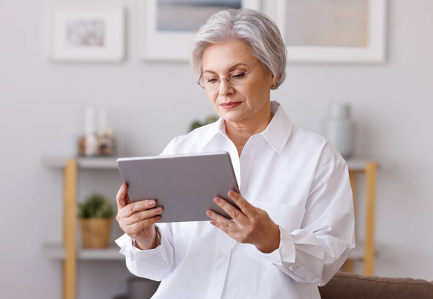 Geschäftsfrau mittleren Alters mit grauen Haaren surft Tablet, während sie zu Hause konzentriert auf den Bildschirm im Wohnzimmer schaut - Foto, Bild