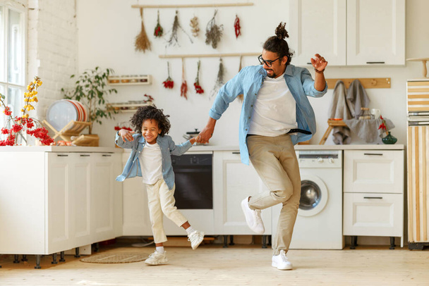 Nadšený šťastný africký americký rodinný otec a malý syn tančí na hudbu v moderní kuchyni, zatímco tráví čas spolu doma, chlapec baví s pozitivním tatínkem o víkendu - Fotografie, Obrázek