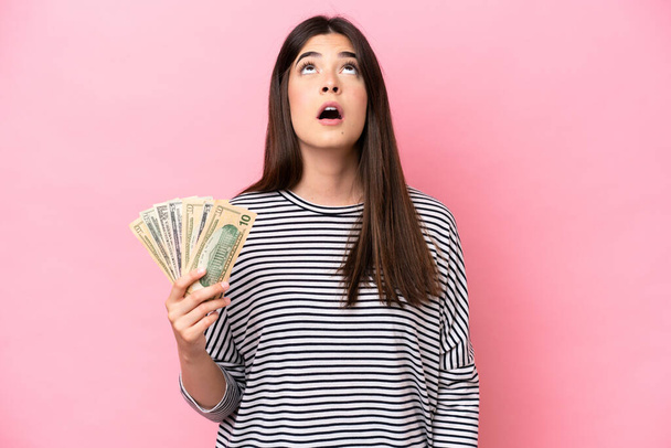 Young Brazilian woman taking a lot of money isolated on pink background looking up and with surprised expression - Photo, Image
