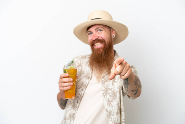 Uomo dai capelli rossi con lunga barba che beve un cocktail su una spiaggia isolata su sfondo bianco che punta davanti con espressione felice - Foto, immagini