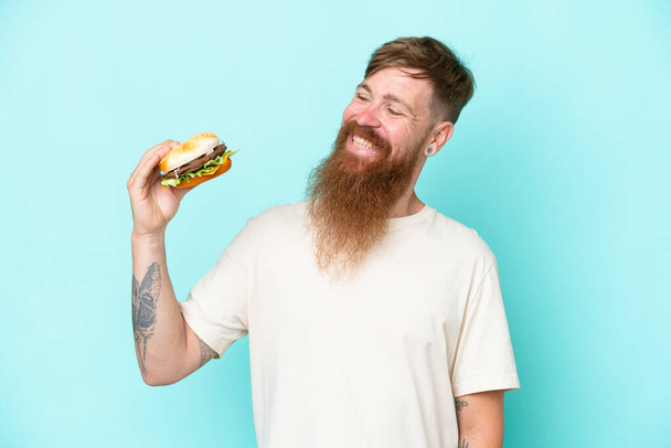 Uzun sakallı kızıl saçlı adam elinde hamburgerle mavi arka planda mutlu bir ifadeyle izole edilmiş. - Fotoğraf, Görsel