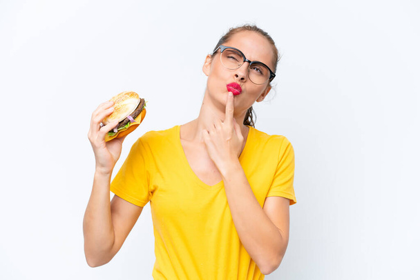 Joven mujer caucásica sosteniendo una hamburguesa aislada sobre fondo blanco teniendo dudas mientras mira hacia arriba - Foto, imagen