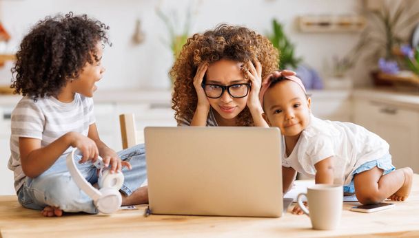 Young irritated stressed African-American mother covering ears and looking at laptop screen while working remotely from home with two small kids, being distracted from work by noisy naughty children - Photo, Image