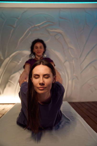 Wellness centrum masażystki podnoszenie tułowia zrelaksowanej młodej kobiety off futon podczas tajskiej sesji masażu - Zdjęcie, obraz