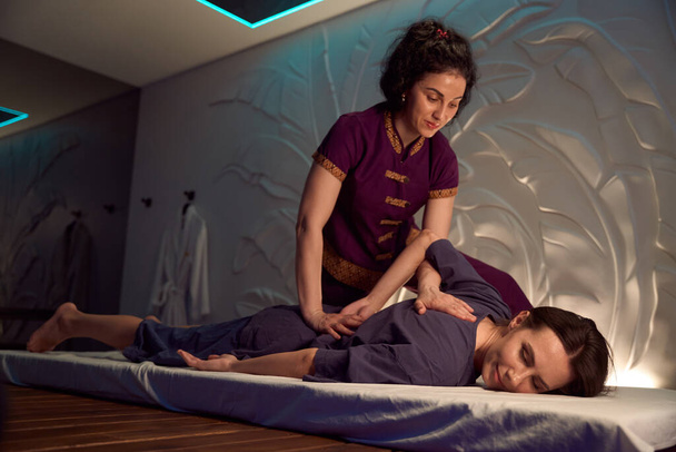 Masoterapeuta kładący ręce nad lewą łopatką i przez staw krzyżowo-biodrowy po prawej stronie pacjenta podczas tajskiej sesji masażu - Zdjęcie, obraz