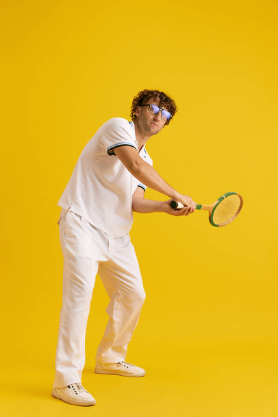 Mature man in glasses and white clothes with tennis racket, training, playing against yellow studio background. Concept of human emotions, facial expression, lifestyle, sport, hobby, ad - Photo, Image