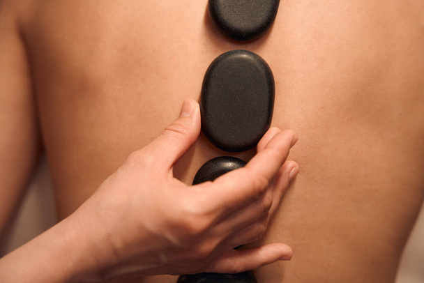 Zbliżenie ręki masażystki umieszczającej płaskie gładkie kamienie bazaltowe wzdłuż kręgosłupa klienta podczas sesji masażerskiej - Zdjęcie, obraz