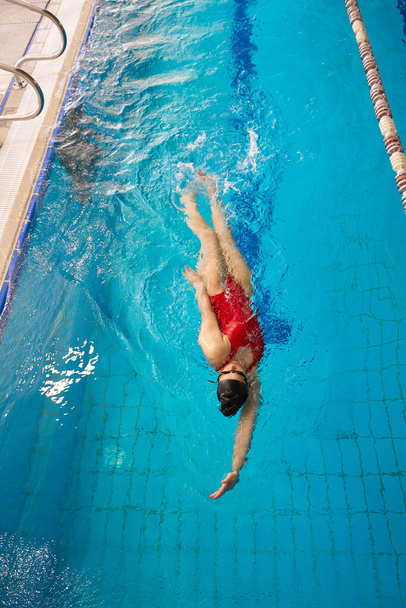Top view of sportswoman in goggles and swim cap practicing backstroke in water - Foto, imagen