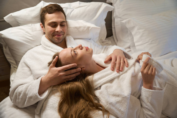 Knappe man knuffelen en strelen aantrekkelijke vriendin die liggen op zijn borst, paar rusten en dromen liggend op bed in badjassen - Foto, afbeelding