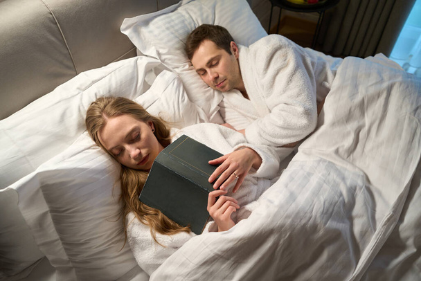 Дружина засинає читаючи книгу, що лежить поруч сплячий чоловік, пара відпочиває після важкого робочого дня в комфортному ліжку з ортопедичними подушками та матрацом - Фото, зображення