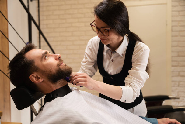 Женщина парикмахер в очках работает с клиентами бородой, мужчина сидит в удобном кресле - Фото, изображение
