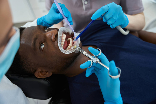 女性の歯科医は男性の顧客に口の引き込み器を挿入し,歯の処置,根管療法または歯の抽出の前に麻酔注入をする - 写真・画像