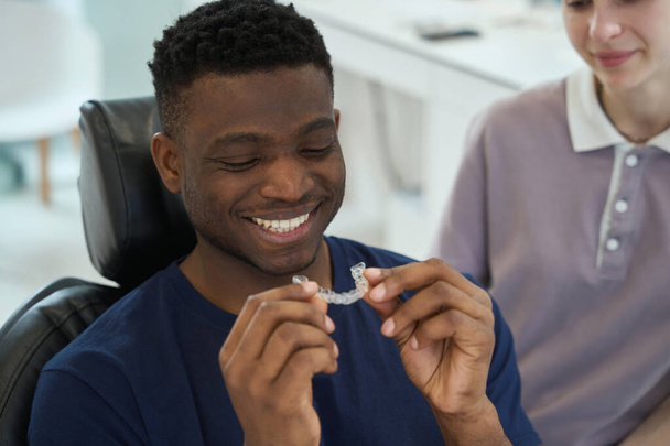 Sonriente hombre afroamericano va a usar frenos claros con el fin de deshacerse de la maloclusión, enfermera cariñosa que le ayuda, odontología estética - Foto, imagen