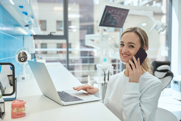 Mujer joven médico utilizando el ordenador portátil y hablando en el teléfono inteligente en el hospital, mirando a la cámara - Foto, imagen