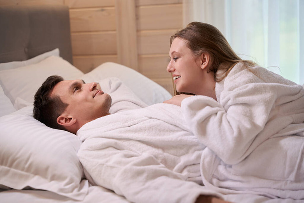 ベッドに滞在中に白いドレッシングガウンを身に着けている幸せな男性に横たわる笑顔の女性 - 写真・画像