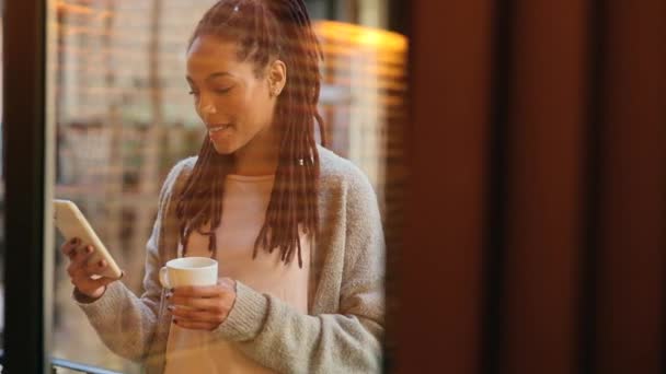 mulher americana africana bonita inclinando-se para fora na varanda em casa com café e telefone inteligente - Filmagem, Vídeo