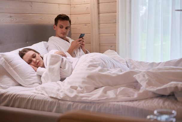 Man houdt smartphone in handen en kijken naar slapende dame terwijl het leggen in bed - Foto, afbeelding