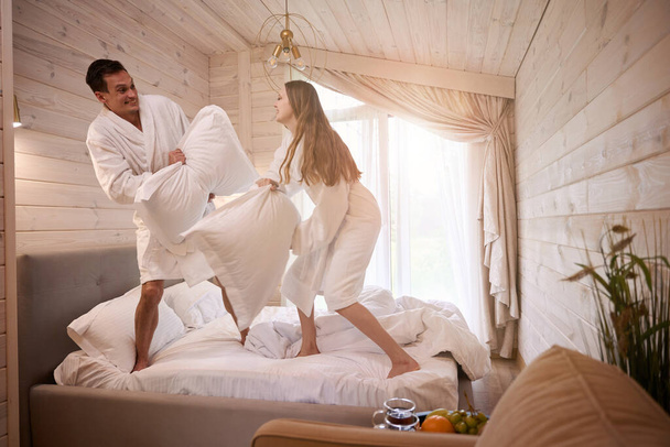 Szczęśliwa para w szlafrokach i zabawy walki na poduszki w sypialni - Zdjęcie, obraz