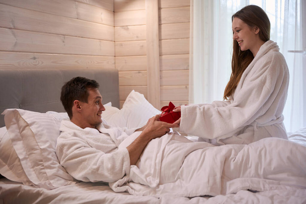 Счастливая дама в халате и дарит подарок улыбающемуся мужчине, лежащему в постели - Фото, изображение