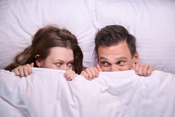 Vista dall'alto di divertenti facce di coppia sdraiato a letto e coperto con coperta bianca - Foto, immagini