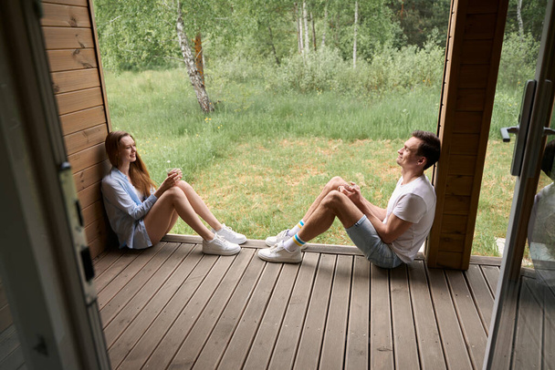 Молода пара сидить на краю котеджної тераси, тримаючи чашки і дивлячись один на одного - Фото, зображення