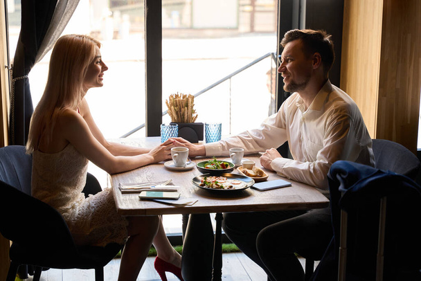 Hermosa pareja teniendo cita en el restaurante, comiendo sabrosa comida y comunicándose sentado en la acogedora mesa, disfrutando el uno del otro - Foto, Imagen