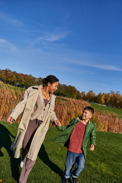 bonding, autumnal nature, happy african american mother holding hands with son, family in outerwear - Fotó, kép