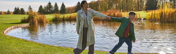 family bonding, happy african american woman following son, walking along pond, hold hands, banner - Foto, Imagem