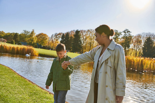 family bonding, cheerful african american mother walking with son along pond, hold hands, autumn - Фото, зображення