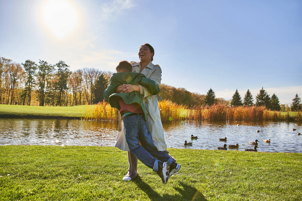 candid, autumn season, happy african american mother lifting playful son, having fun near pond - Foto, afbeelding