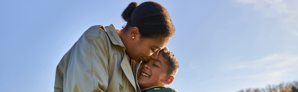 candida, stagione autunnale, felice donna afroamericana in capispalla abbracciando allegro figlio, striscione - Foto, immagini