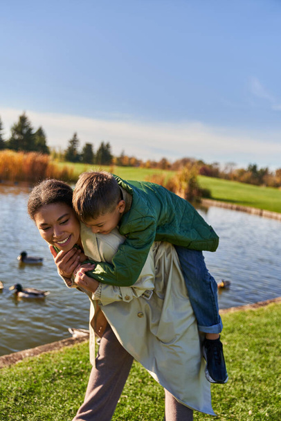 fall colors, happy african american woman piggybacking son near pond with ducks, childhood, autumn - Foto, Imagem