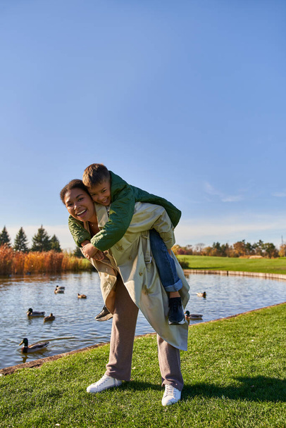 fall colors, joyful african american woman piggybacking boy near pond with ducks, childhood, autumn - Valokuva, kuva