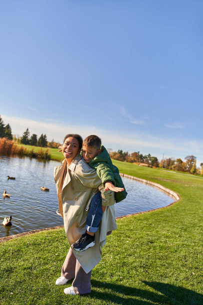 fall colors, mother piggybacking son near pond with ducks, happy childhood, african american, autumn - Fotó, kép