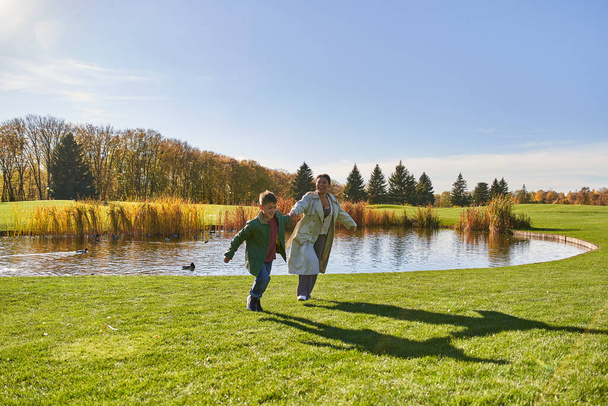 playful, happy african american mother and son running on grass near pond, modern parenting, fun - Foto, afbeelding