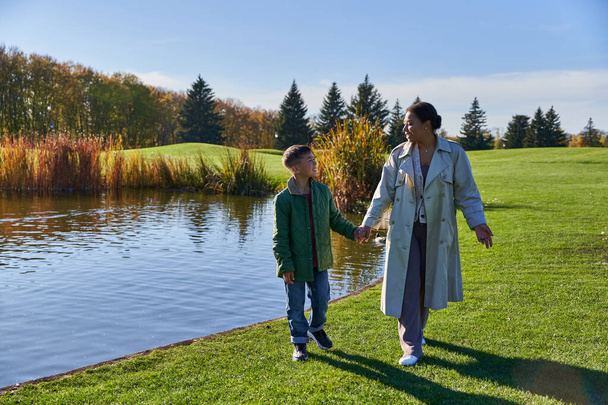 mother and son holding hands and walking on grass near lake, african american family, tranquil - Foto, afbeelding