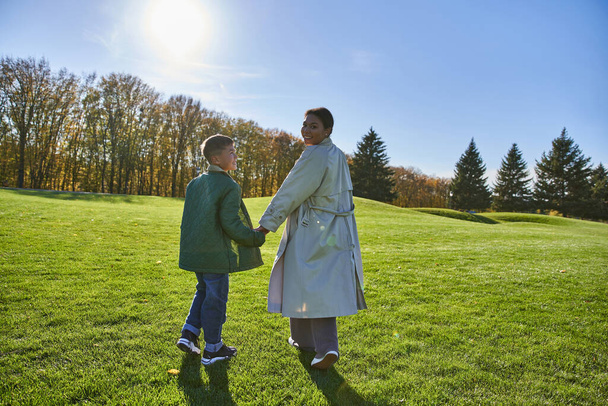 sunny day in fall, african american woman walking with son in park, green grass, autumn, outerwear - Foto, Bild