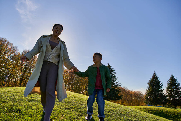 vidám afro-amerikai anya és gyermek kéz a kézben, séta le a dombról, őszi ruhák - Fotó, kép