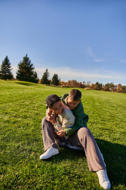 Χαρούμενη μητέρα και γιος κάθονται στο γρασίδι, ηλιόλουστη μέρα, φθινόπωρο, παιχνιδιάρικο αφροαμερικάνικο αγόρι αγκαλιάζει τη μαμά - Φωτογραφία, εικόνα