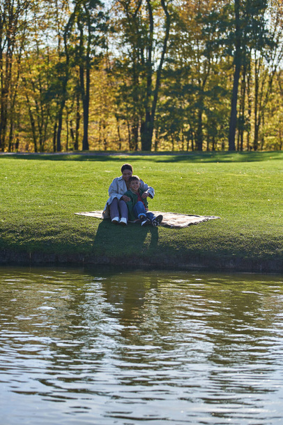 cheerful mother and son hugging in autumnal park, sitting on blanket near lake, african american - Photo, Image