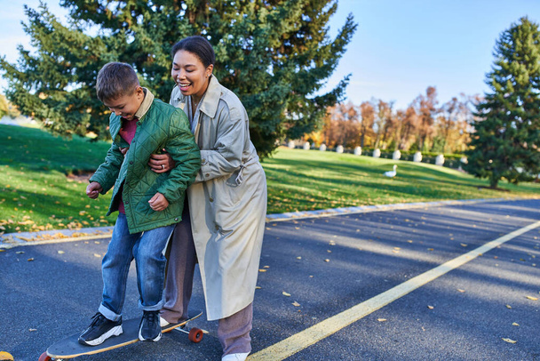happy mother and son in autumn park, african american woman teaching boy how to ride penny board - Foto, Imagem