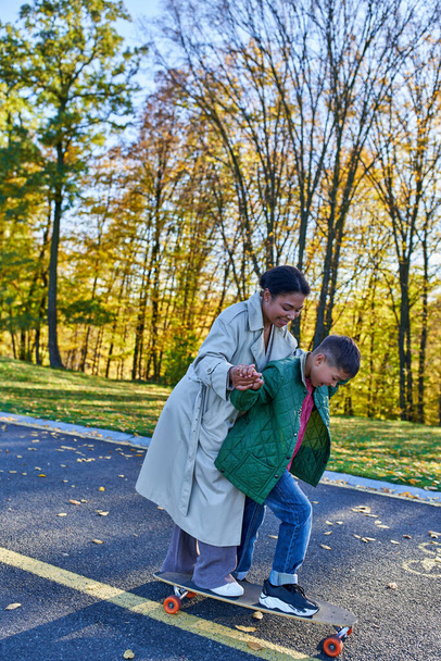 mother and son, bonding time, autumn park, african american woman walking near boy on penny board - Фото, зображення